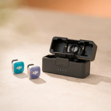 ಗ್ಯಾಲರಿ ವೀಕ್ಷಕಕ್ಕೆ ಚಿತ್ರವನ್ನು ಲೋಡ್ ಮಾಡಿ, DJI Mic Mini 2S transmitters with teal and purple magnetic covers on a sandy surface, next to an open black charging case holding the receiver, with blue charge indicator lights lit.