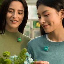 गैलरी व्यूवर में इमेज लोड करें, Two smiling women in green and teal sweaters looking at flowers, each wearing a small green DJI Mic Mini 2 transmitter on her chest.
