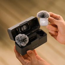गैलरी व्यूवर में इमेज लोड करें, Two hands hold an open DJI Mic Mini 2 charging case with a transmitter inside wearing a fluffy windscreen, while the other hand holds up a white transmitter with a windscreen.
